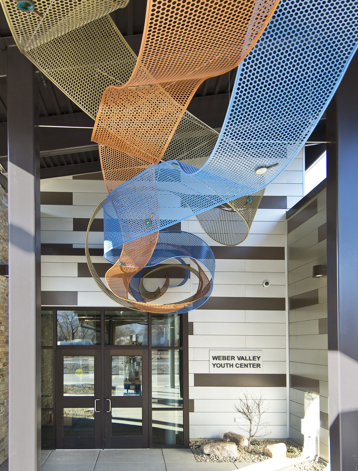 A image of an outdoor, metal sculpture resembling yellow, orange, and blue ribbons.