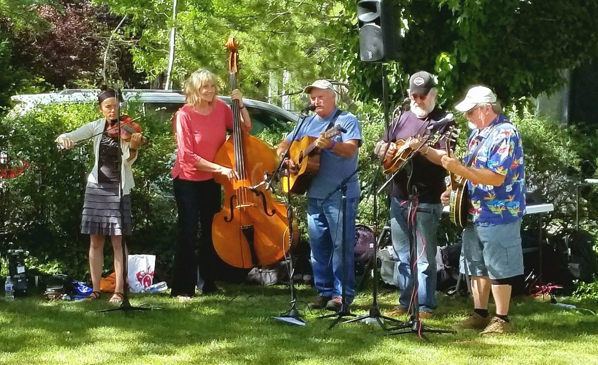 A group of five musicians plays music in a green, leafy outdoor setting.
