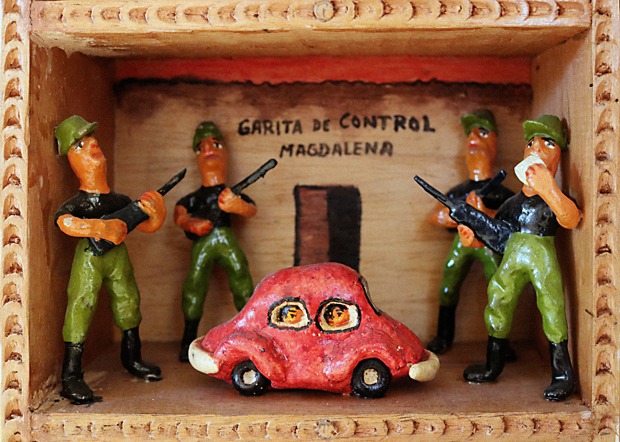 A scene called Magdalena Control Point within a retablo. The scene portray four large figurines dressed in military-like uniforms and holding guns, towering over a red car. In the car windows can be seen faces of passengers. The back wall has text that reads, "Garita de Control Magdalena."