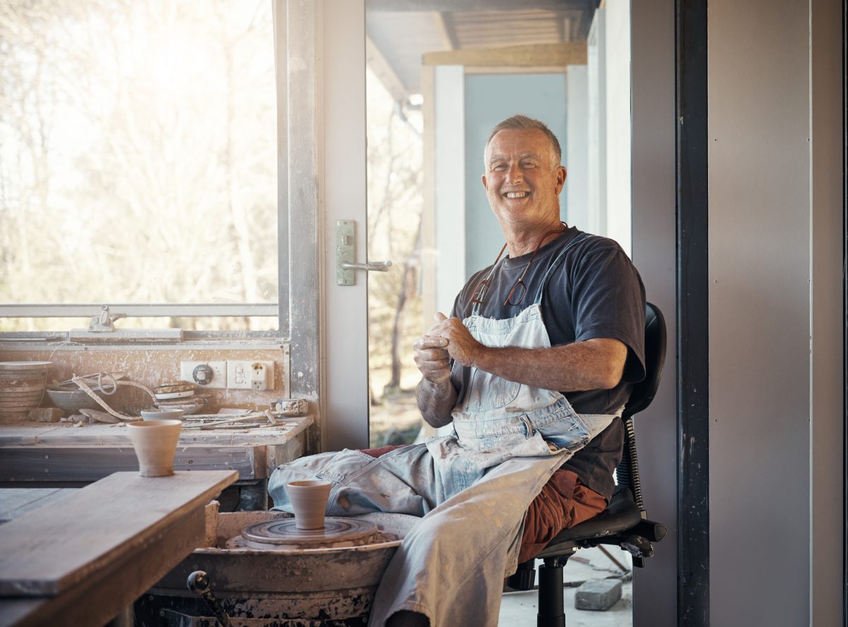 A white man wearing a dark grey shirt and white apron with short blond hair sits making pottery.