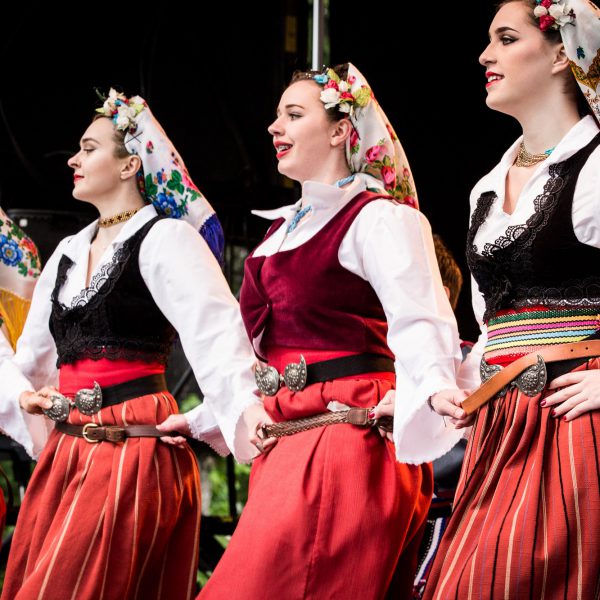 Three women perform a folk dance wearing white blouses, red skirts, and colorful headscarves.