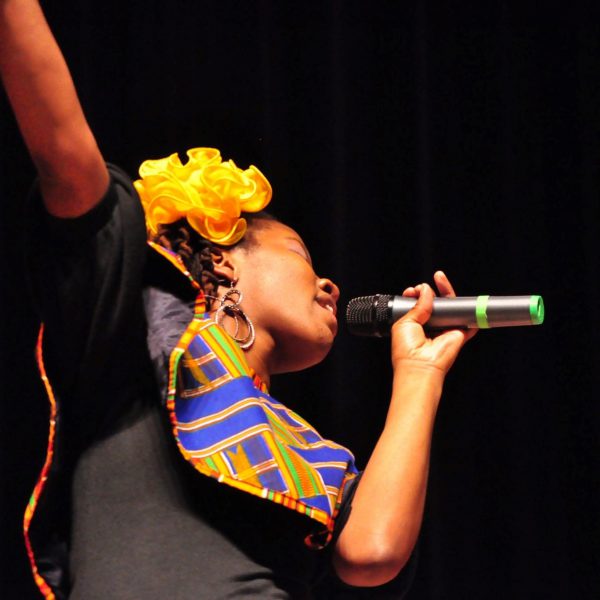 A photo of a woman singing into a microphone with a yellow flower in her hair.