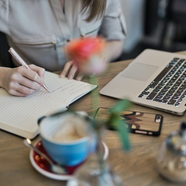 A woman writes in a notebook at a table with a laptop, phone, coffee, and a flower nearby.