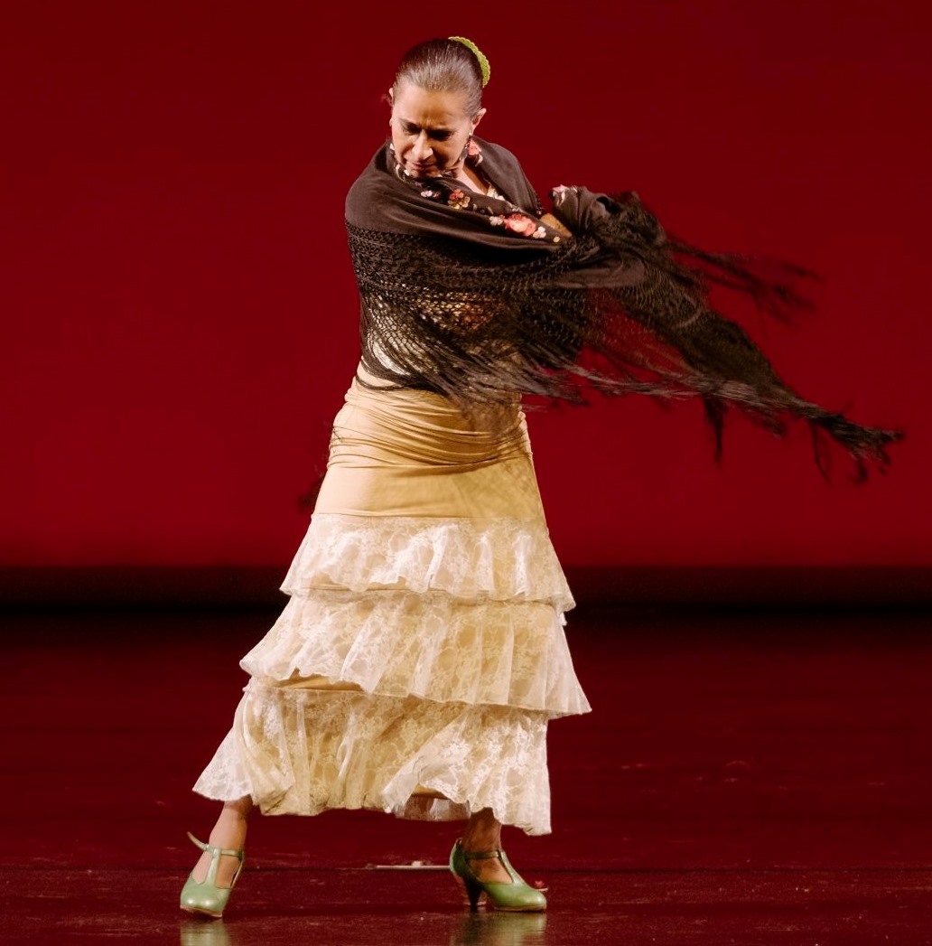 A photo of a woman wearing a white ruffled dress and a brown shawl dancing on a stage with a red background.