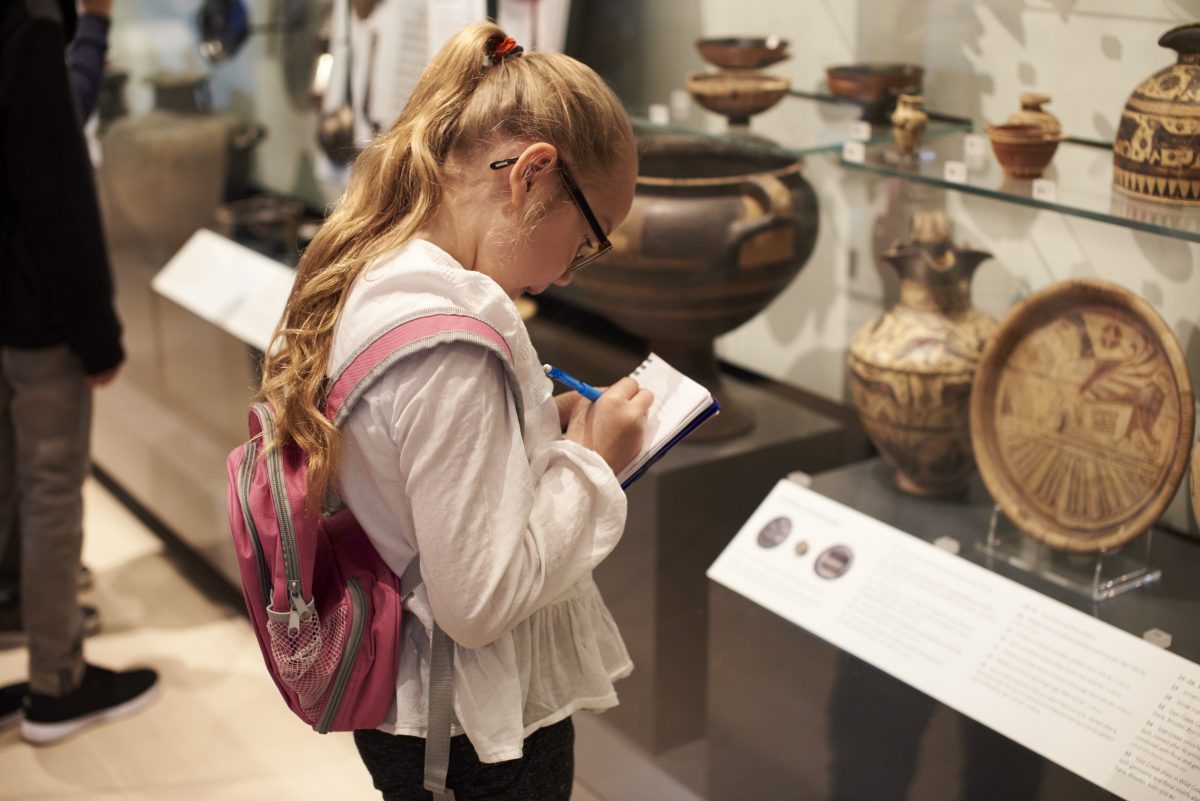 A child with long blonde hair wearing a pink backpack, writing in a notebook in a museum.