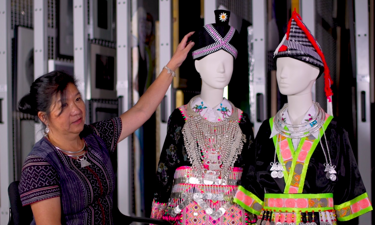 A woman with dark hair sits beside two mannequins wearing traditional Hmong clothing.