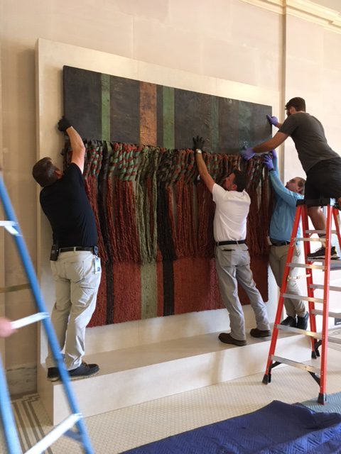 People hang a fabric artwork on a marble wall.