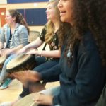 Three female students actively hitting drums while seated in a circle.