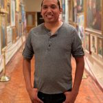 A smiling individual stands in a corridor lined with portraits, wearing a gray long-sleeve shirt and black pants.