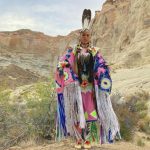An individual in Native American garb standing in front of a scenic desert rock landscape.