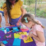 A woman and a young girl engaged in a craft activity at a table. The girl is applying glue to a butterfly-shaped cutout while the woman observes. The table is decorated with colorful paper, scissors, and craft supplies.