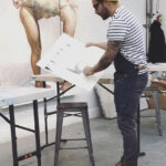 A white male in black pants, striped shirt, yellow beanie, and brown work boots stands in an art studio sketching shapes on a large white paper. A painting behind him on the wall shows a nude female form.