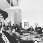 a black and white photo of author Jared Quan in a conference foom with others seated at a table behind him