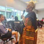 A woman in a brightly patterned traditional African garment speaks into a microphone while standing in front of an audience. In the foreground, a young person with curly hair sits in a chair, wearing a striped shirt and shorts.