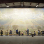 A wall-size mural of the sun breaking through clouds with multiple observers standing in front