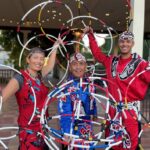Three individuals performing a hoop dance pose with colorful hoops, wearing traditional attire and headdresses, in a vibrant outdoor setting.
