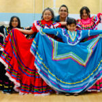 Four dance students in colorful latin dance dresses pose with teaching artist Raquel Baez