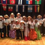 a croup of fifteen children in dance latin dance costumes pose beneatth rows of international flags