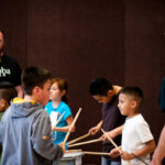 Children using drumsticks pound on drums, as Samba Fogo musician with whistle in his mouth looks on