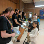 Drummers holding large drums stand in a line while young students play the drums