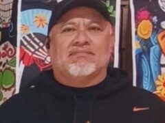 **Alt Text:** A man with a serious expression stands in front of colorful Day of the Dead-themed backdrop featuring skulls and floral designs.
