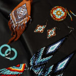 A collection of handmade beaded jewelry displayed on a black fabric background. The assortment includes various earrings, a necklace, and a pendant with intricate beadwork in vibrant colors.