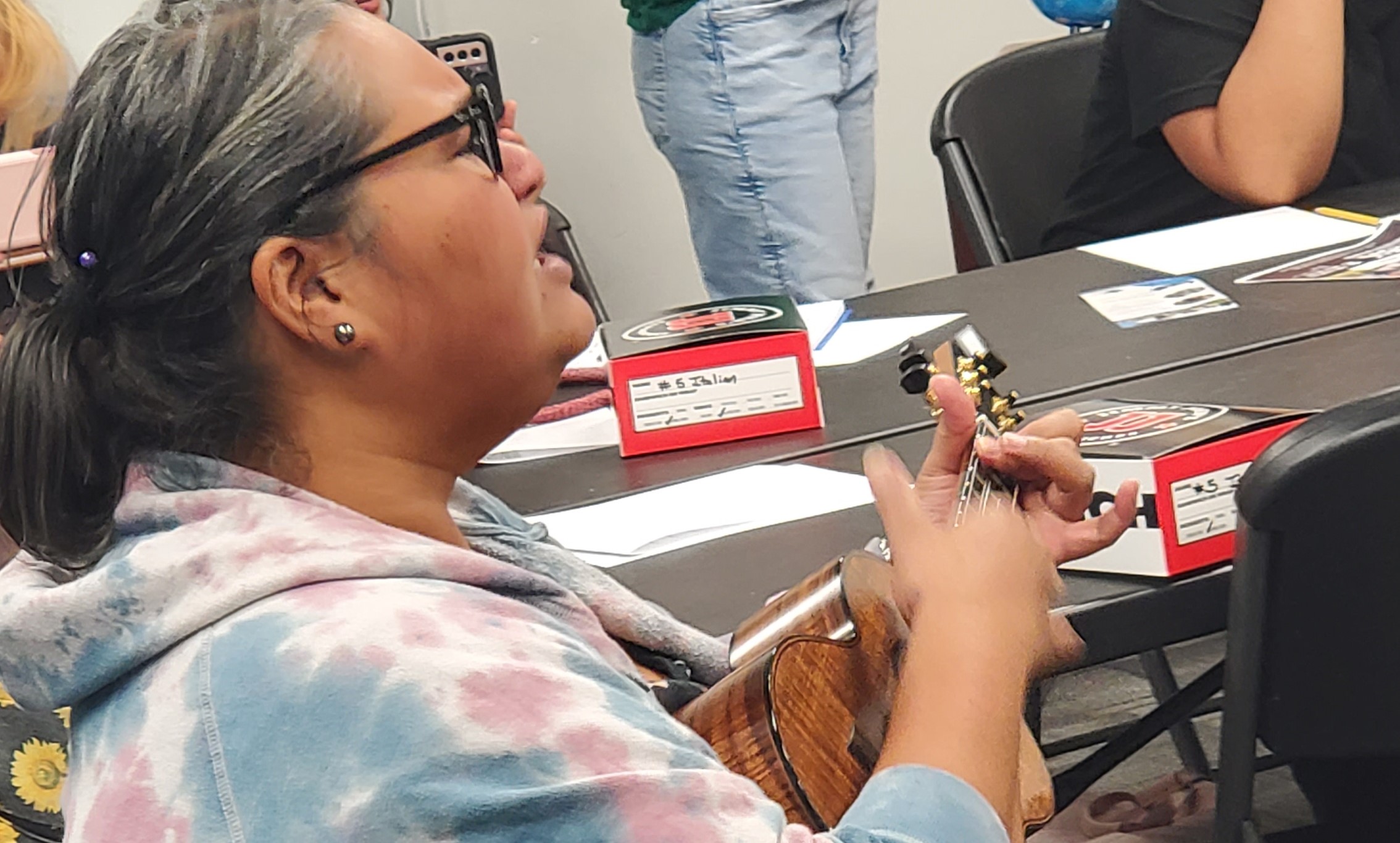 A person with shoulder-length hair and glasses is sitting in a classroom, strumming a ukulele. They are wearing a tie-dye hoodie and a floral-patterned dress. In the background, other individuals are holding phones and observing.