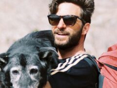 A man standing on a rocky trail in a mountainous area, smiling while holding a black dog in his arms. He is wearing sunglasses, a black shirt with white stripes, and light-colored pants. A large red backpack is secured to his back.