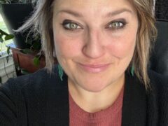 A woman with short, tousled hair smiles at the camera. She is wearing a deep red sweater under a black cardigan, and her earrings are small and turquoise. In the background, there are plants and a window allowing natural light into the room.