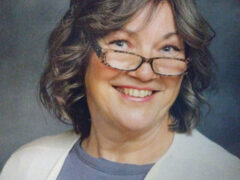 A smiling middle-aged woman with wavy gray hair, wearing glasses and a light beige cardigan over a gray shirt, set against a dark background.
