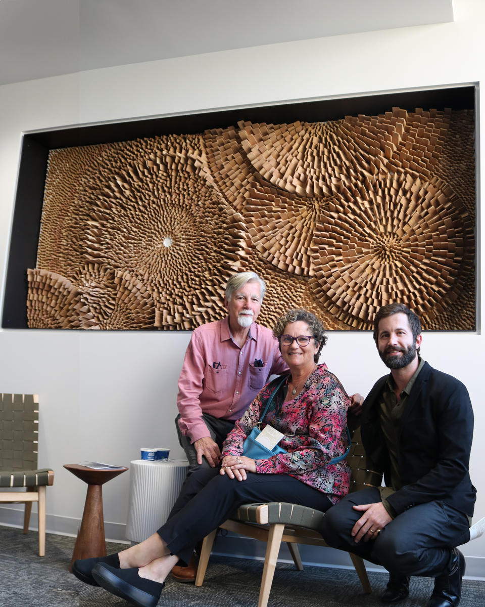 An older couple and their son in front of an intricate wood artwork.