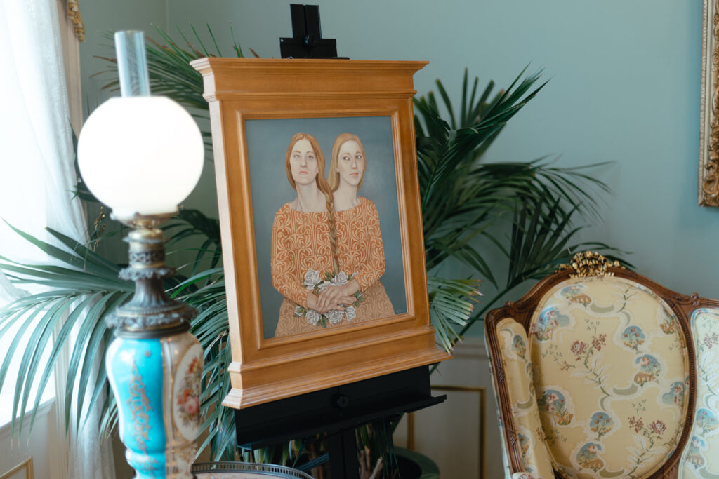 A painting of two women with their long hair braided together stands on an easel in a formal parlor.