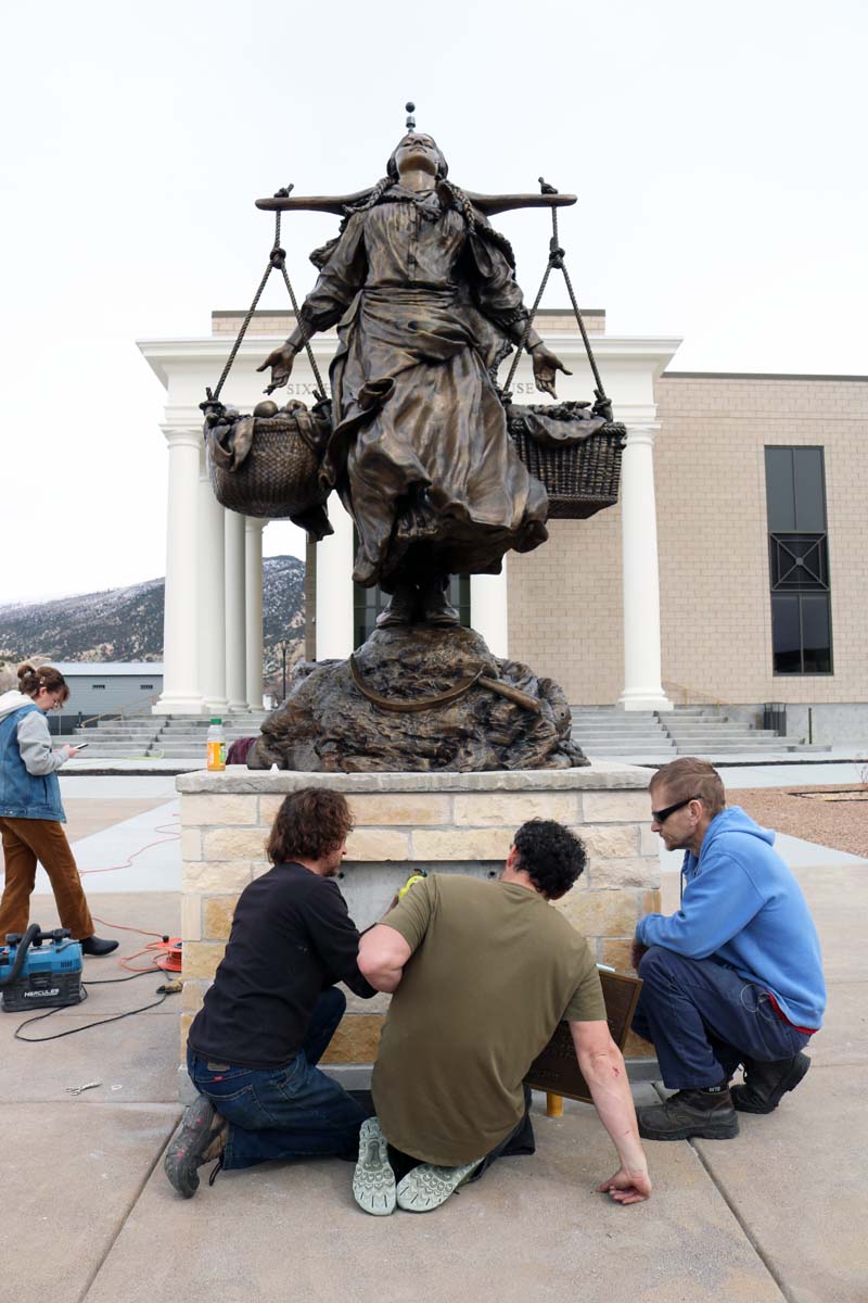 Several men installing a plaque at the base of a large bronze statue of a woman with two baskets of fruit.