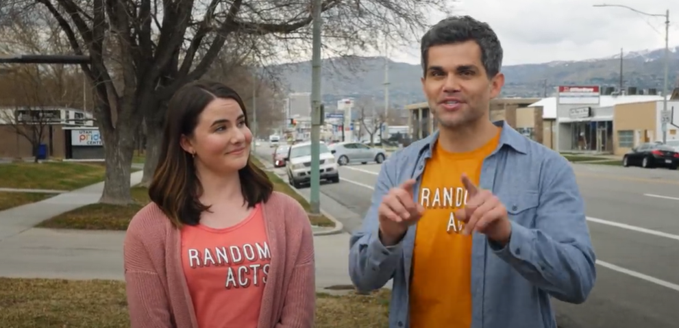 A woman listens while a man speaks to the camera. Both are wearing T-shirts that read "Random Acts."