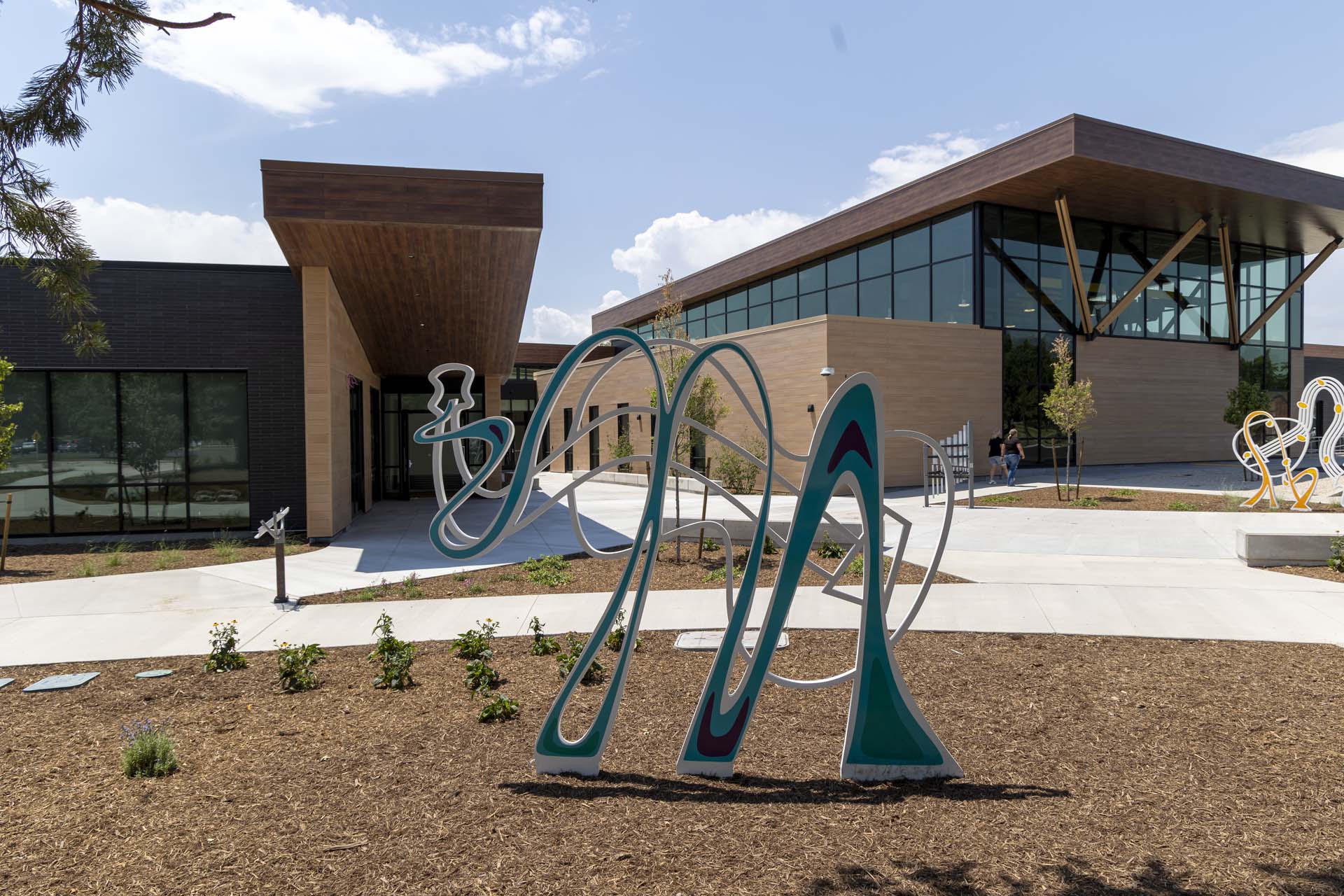 Modern building with large windows and angular wooden roof. Featured in front is a whimsical, looping metal sculpture on a landscaped walkway that resembles an elephant morphed with a guitar. Bright day.