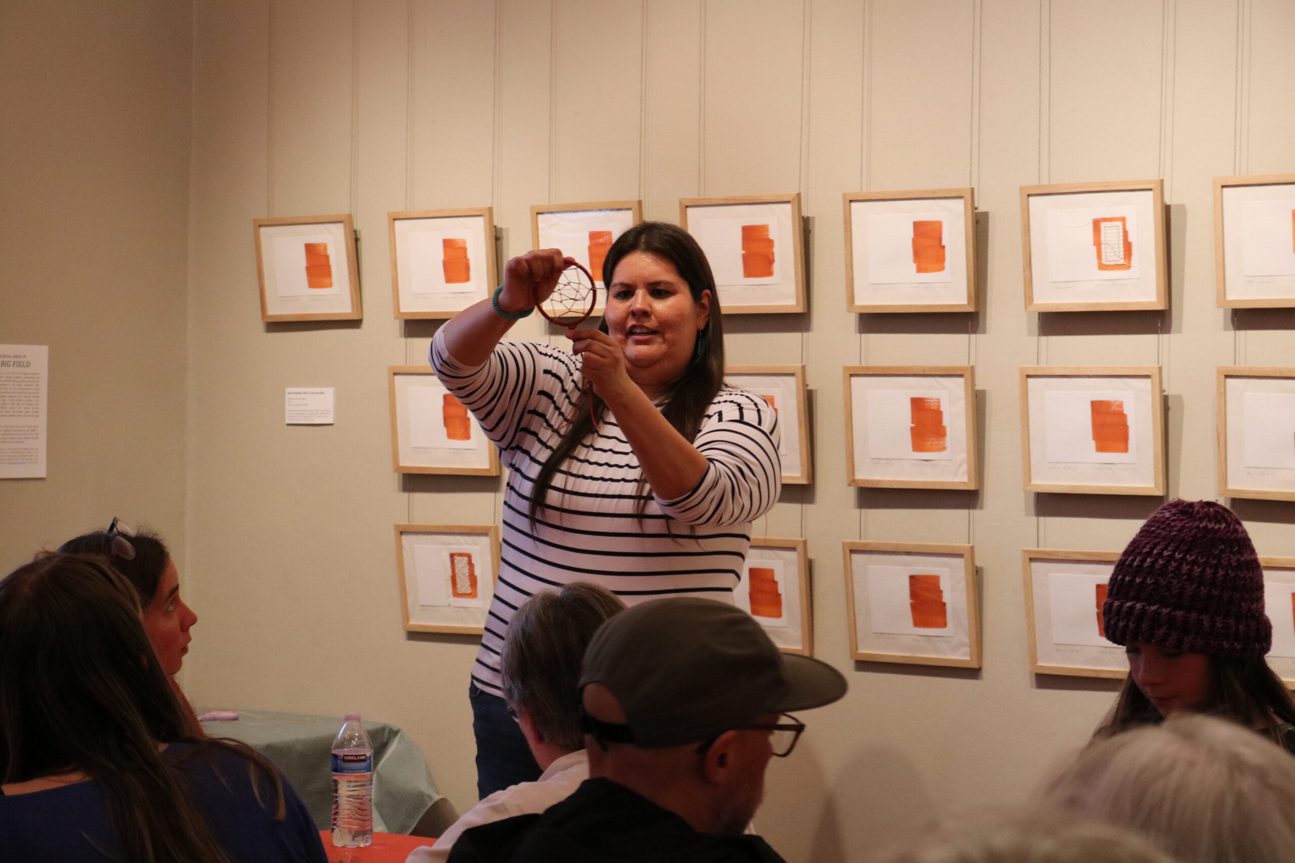 A woman in a striped shirt is demonstrating a craft to a seated audience in an art gallery. The walls display framed abstract orange artworks. The atmosphere is educational and engaged.