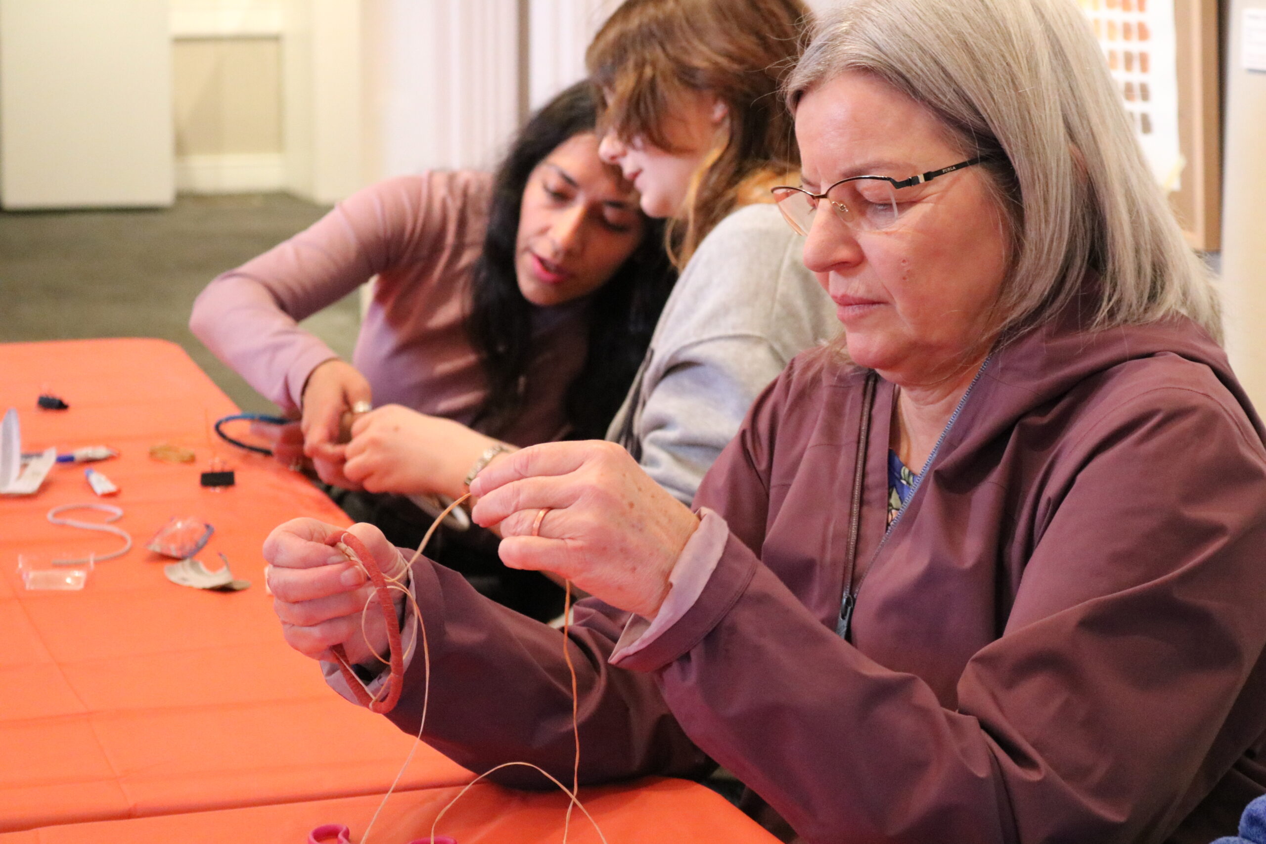 Three women are seated at a table engaged in crafting activities. The woman in front holds strings intently, while two others focus on their project, creating a calm and collaborative atmosphere.