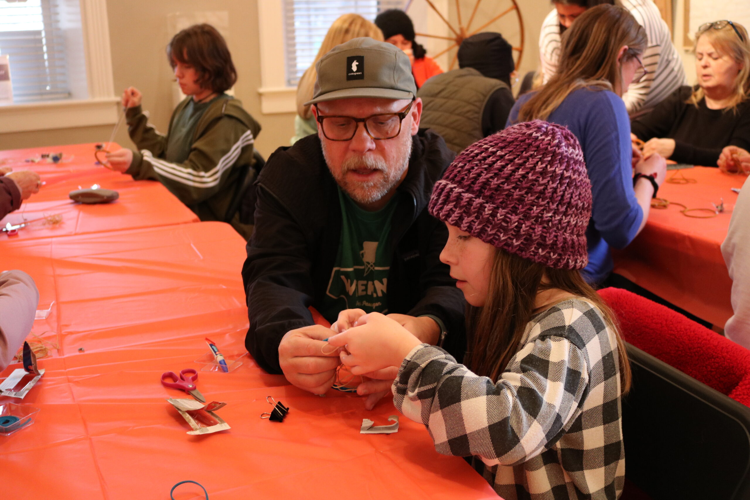 A man in a cap and glasses helps a young girl with a craft project at a table covered with an orange cloth. Others engage in similar activities in the background.