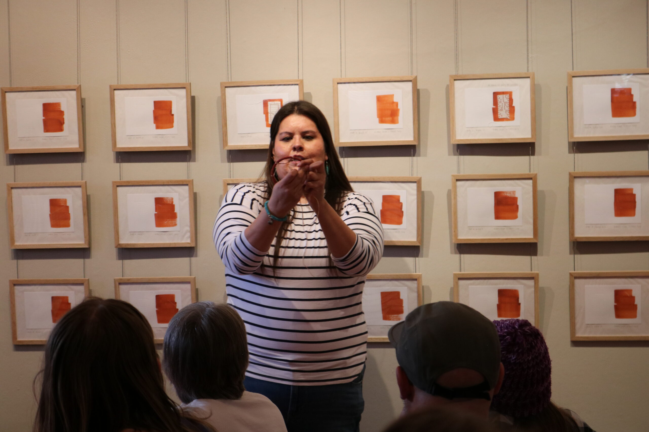 A woman in a striped shirt demonstrates a craft to a seated audience in an art gallery. Framed abstract art pieces with orange elements adorn the wall.