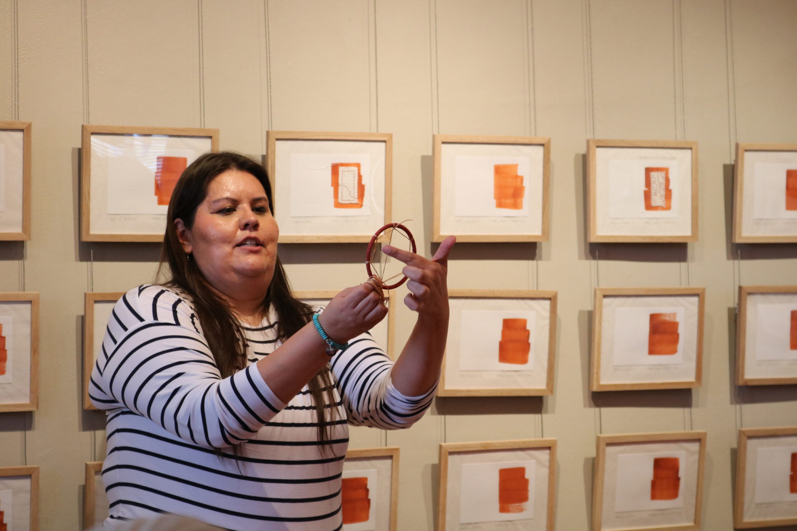 A woman in a striped shirt demonstrates the techniques of weaving a dreamcatcher in an art gallery. Framed orange abstract art hangs in a grid on the wall behind her.