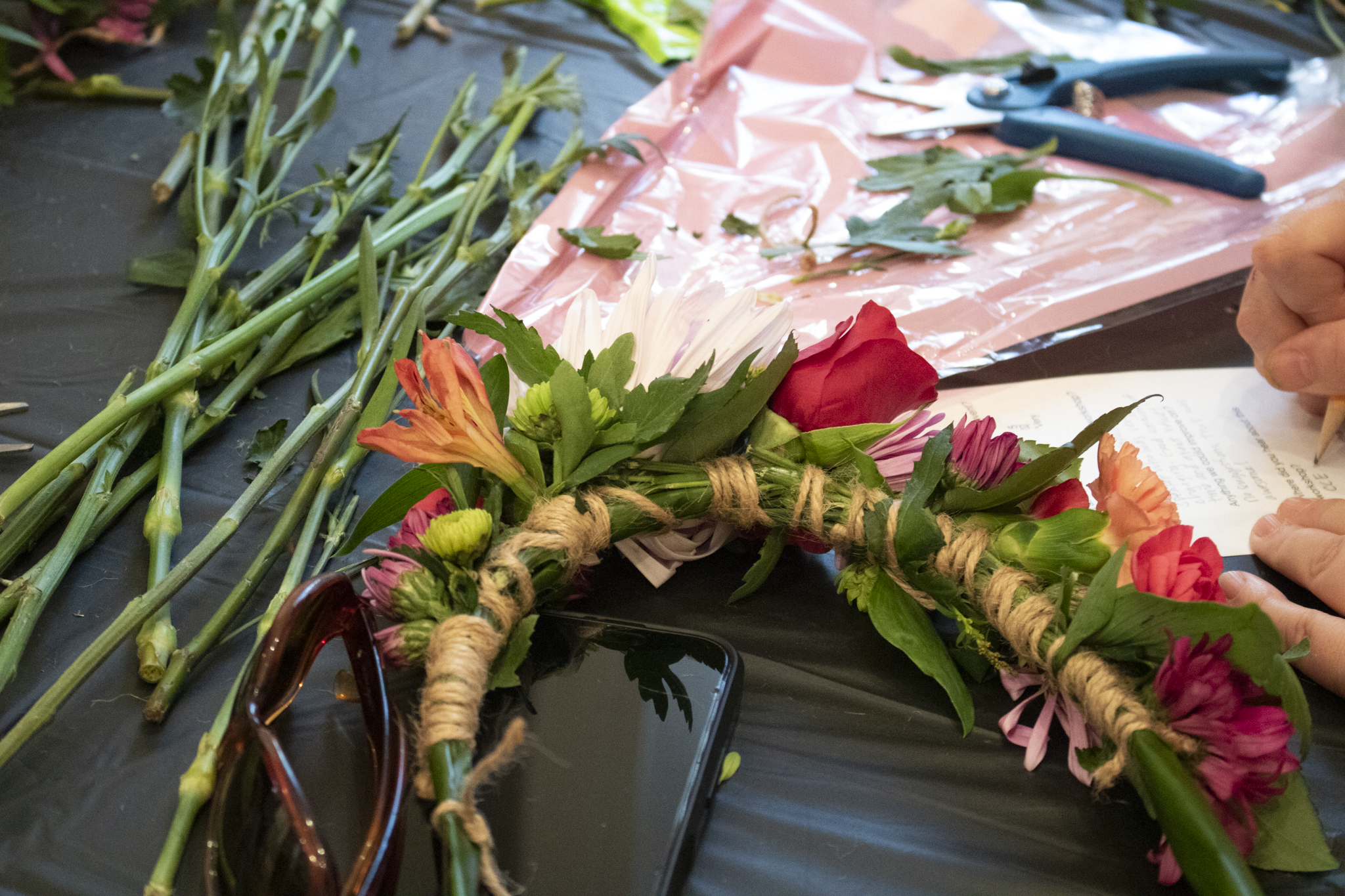 A floral wreath in progress lies on a black table with scattered stems. Next to it are sunglasses and a phone, conveying a creative atmosphere.