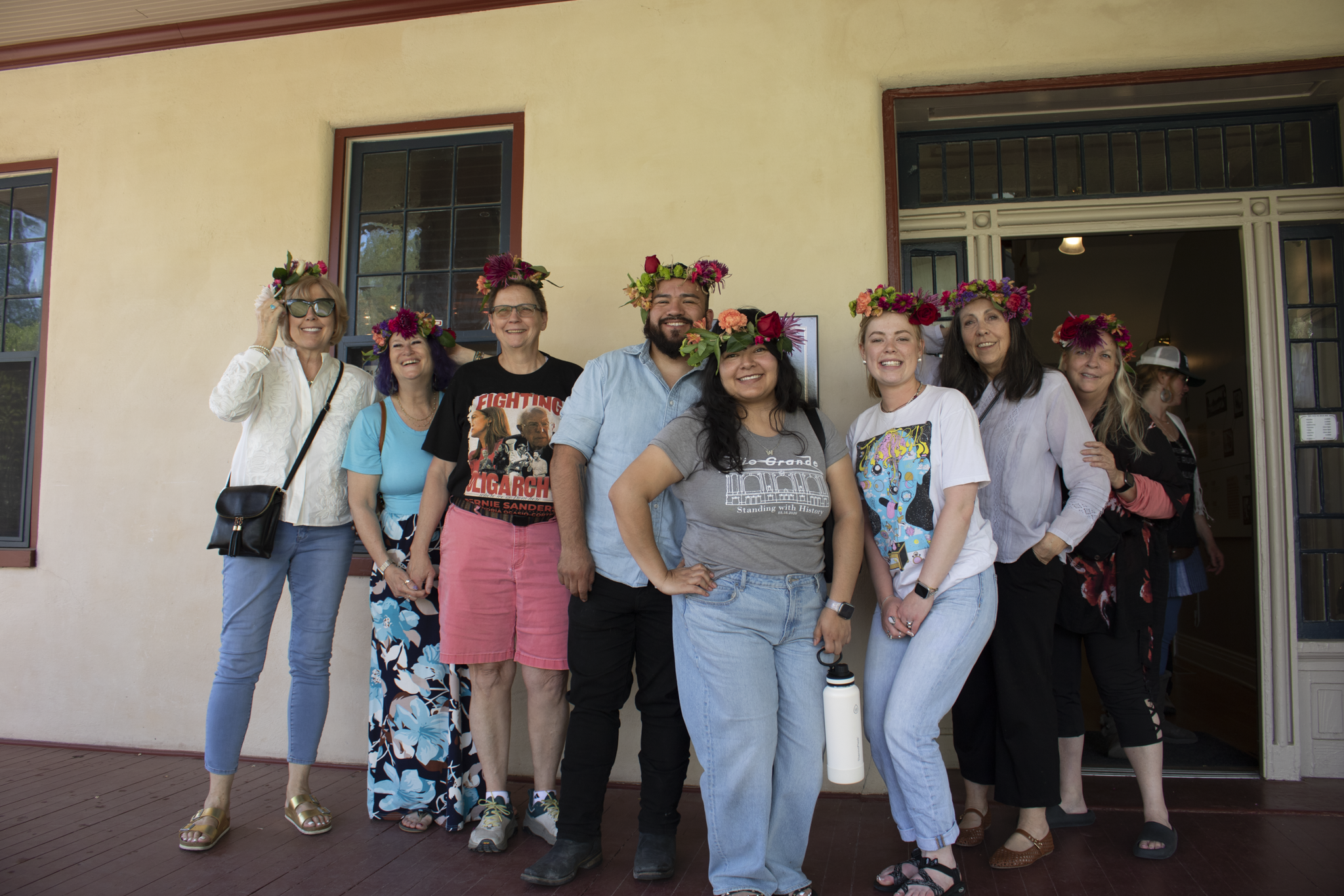 A group of eight people stand smiling on a porch, wearing vibrant flower crowns. The mood is joyful and relaxed, with a warm, sunny-day ambiance.