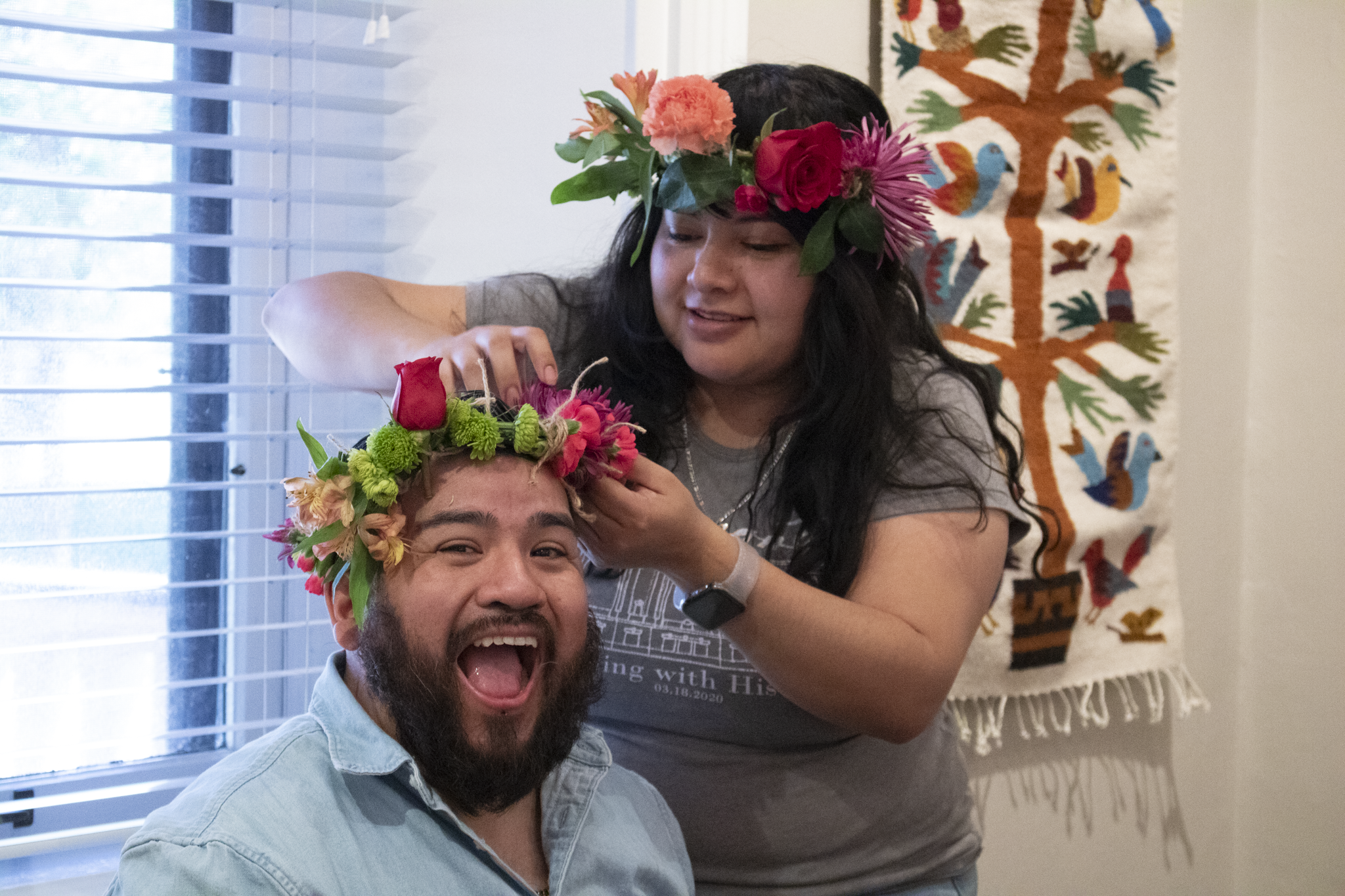 A woman playfully adjusts a colorful flower crown on a smiling man's head. Both wear flower crowns. A vibrant, embroidered wall hanging is in the background.