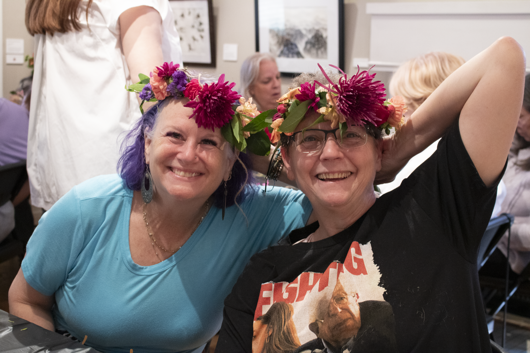 Two smiling individuals wearing flower crowns sit closely. One wears a light blue shirt, the other a black shirt. The setting is a lively gathering.