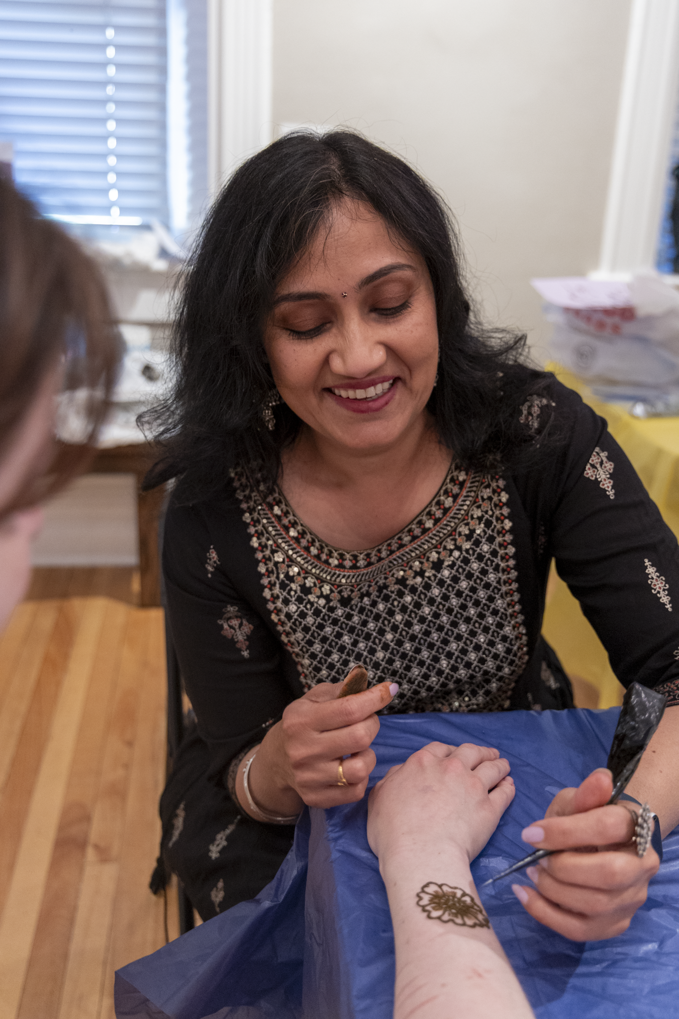 A woman smiles warmly while applying intricate henna designs to another person's forearm. The setting is indoors with wooden floors, creating a cozy vibe.