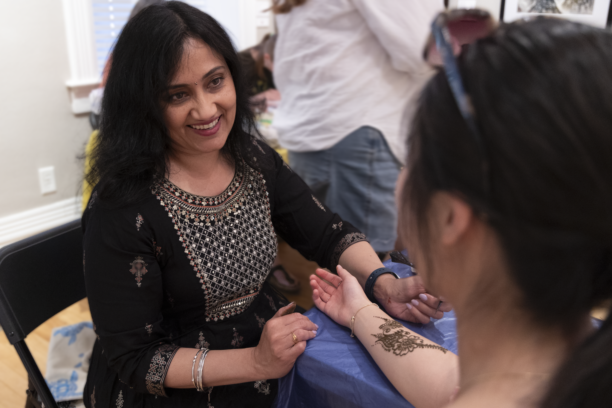 A woman in a black embroidered dress smiles warmly while applying intricate henna designs on another woman’s forearm. The scene conveys creativity and joy.
