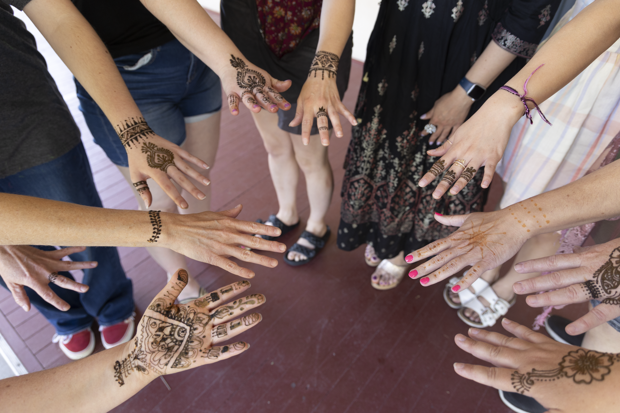 A group of people stand in a circle, showcasing intricate henna designs on their hands. The mood is festive, with diverse outfits visible.
