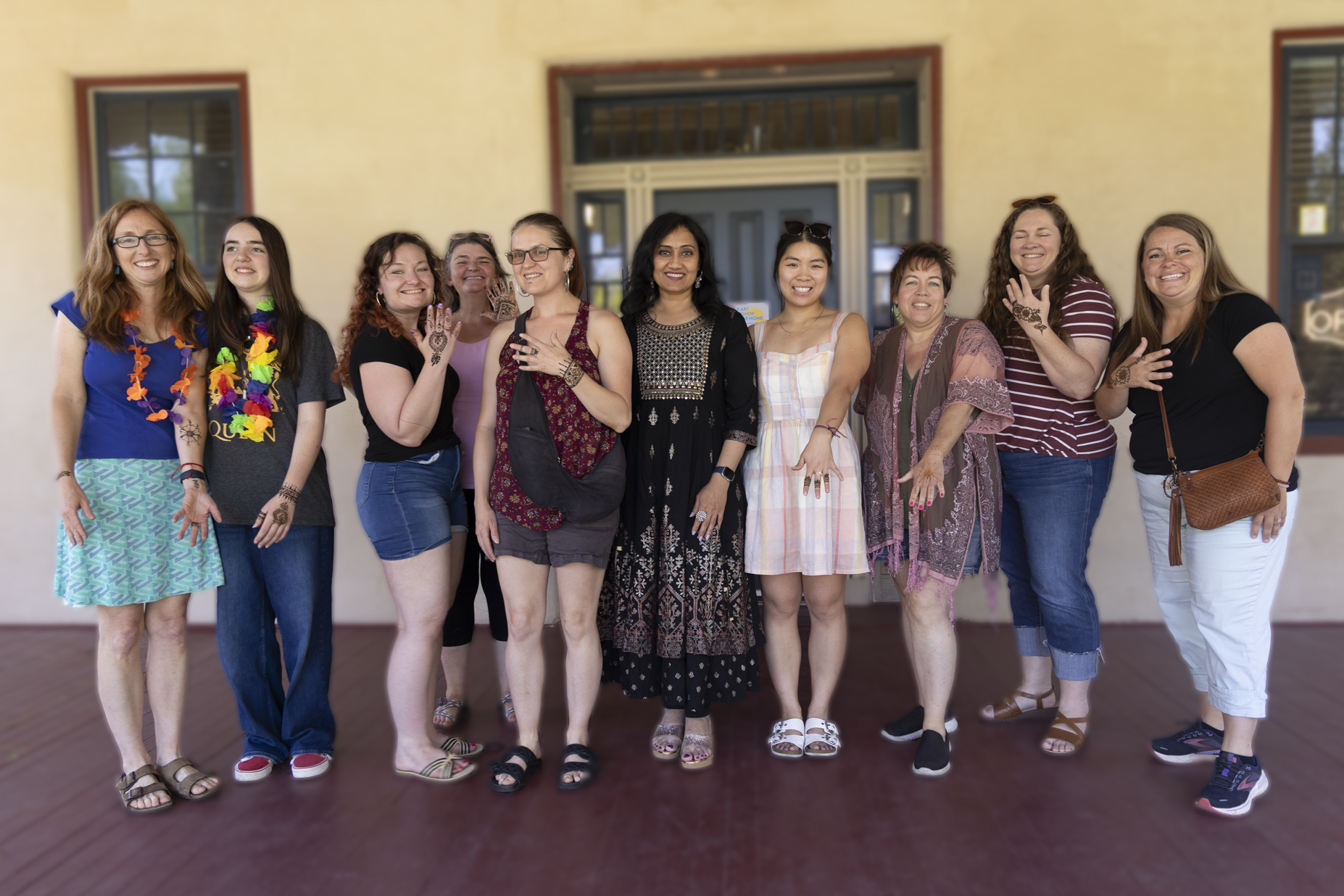 A group of 10 women stand in a line, smiling and showing intricate henna designs on their hands. They are outdoors on a sunny day, conveying happiness.
