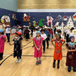 A group of children stand in lines in a gymnasium decorated with winter holiday imagery, including Santa, reindeer, and snowmen on a blue backdrop. Some children wear colorful traditional dresses and headscarves, while others wear white dresses or shirts with cowboy hats.
