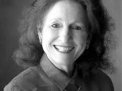 A smiling woman with wavy hair and earrings, wearing a collared shirt and a necklace.